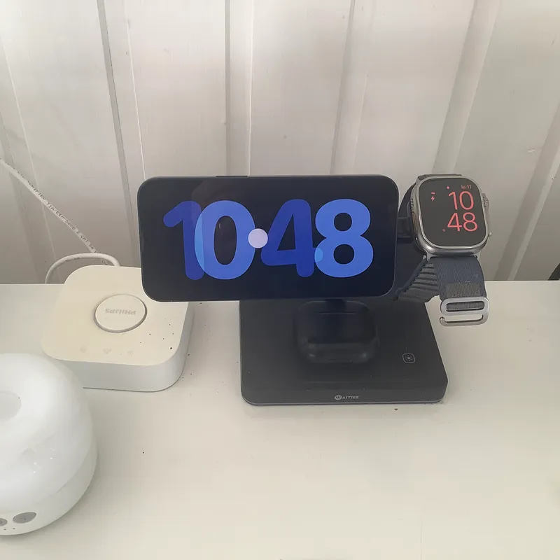 Pöytä, jossa on digitaalinen kello, joka näyttää kellonajan 10:48, valkoinen laite ja älykello.